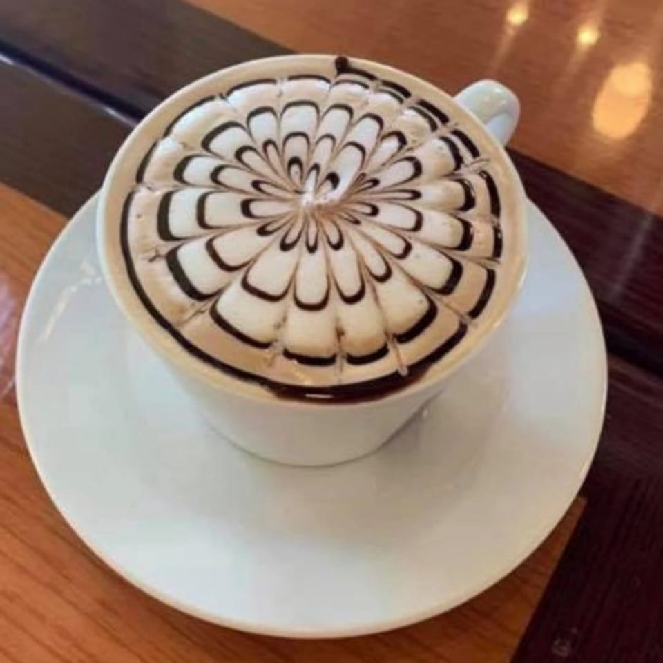 A latte with intricate chocolate drizzle art on top, served in a white cup and saucer.
