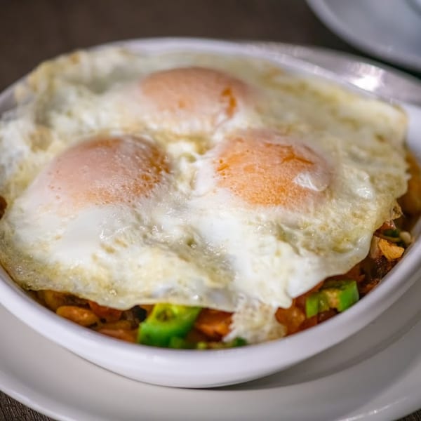 Three fried eggs with runny yolks on a bed of breakfast hash with avocado in a white bowl.