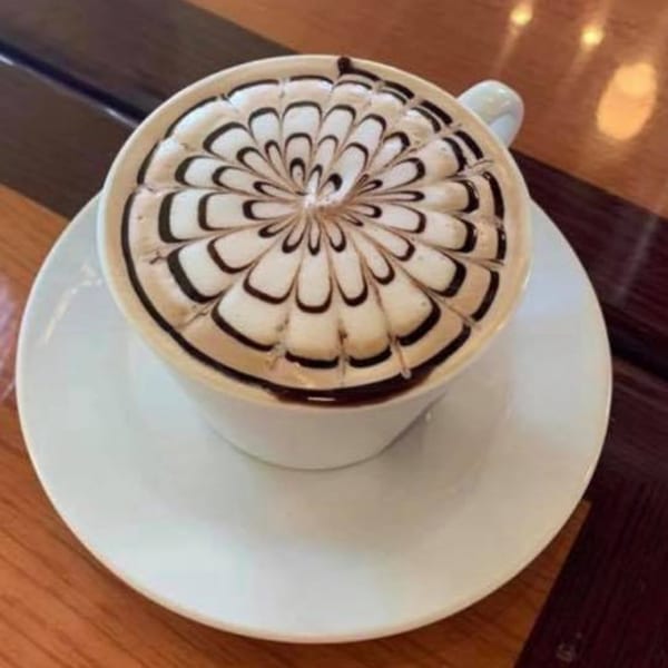 A white cup of coffee with intricate chocolate latte art on a wooden table at Garden Berry Cafe.