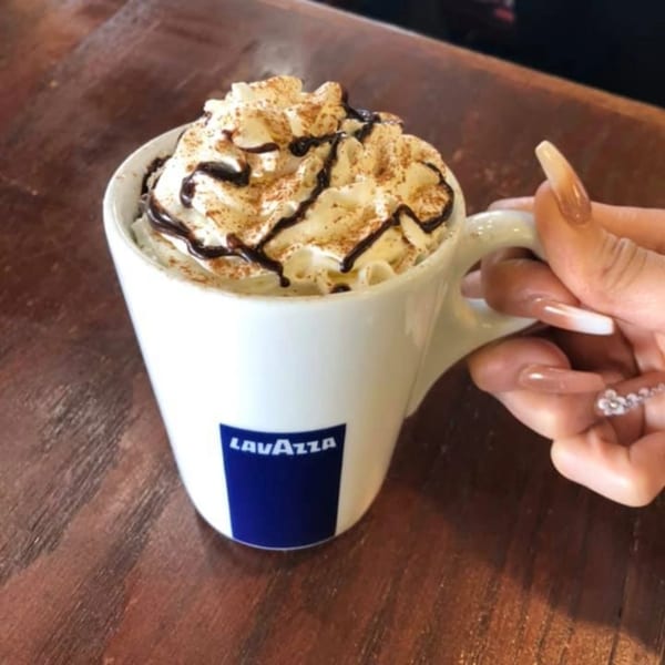 A hand holds a white Lavazza mug filled with a drink topped with whipped cream and chocolate drizzle.